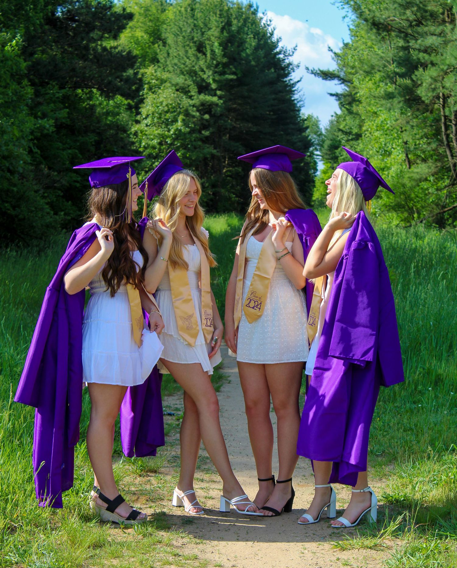 Group photo showing graduates in purple caps and gowns posing playfully on a dirt path surrounded by green trees.