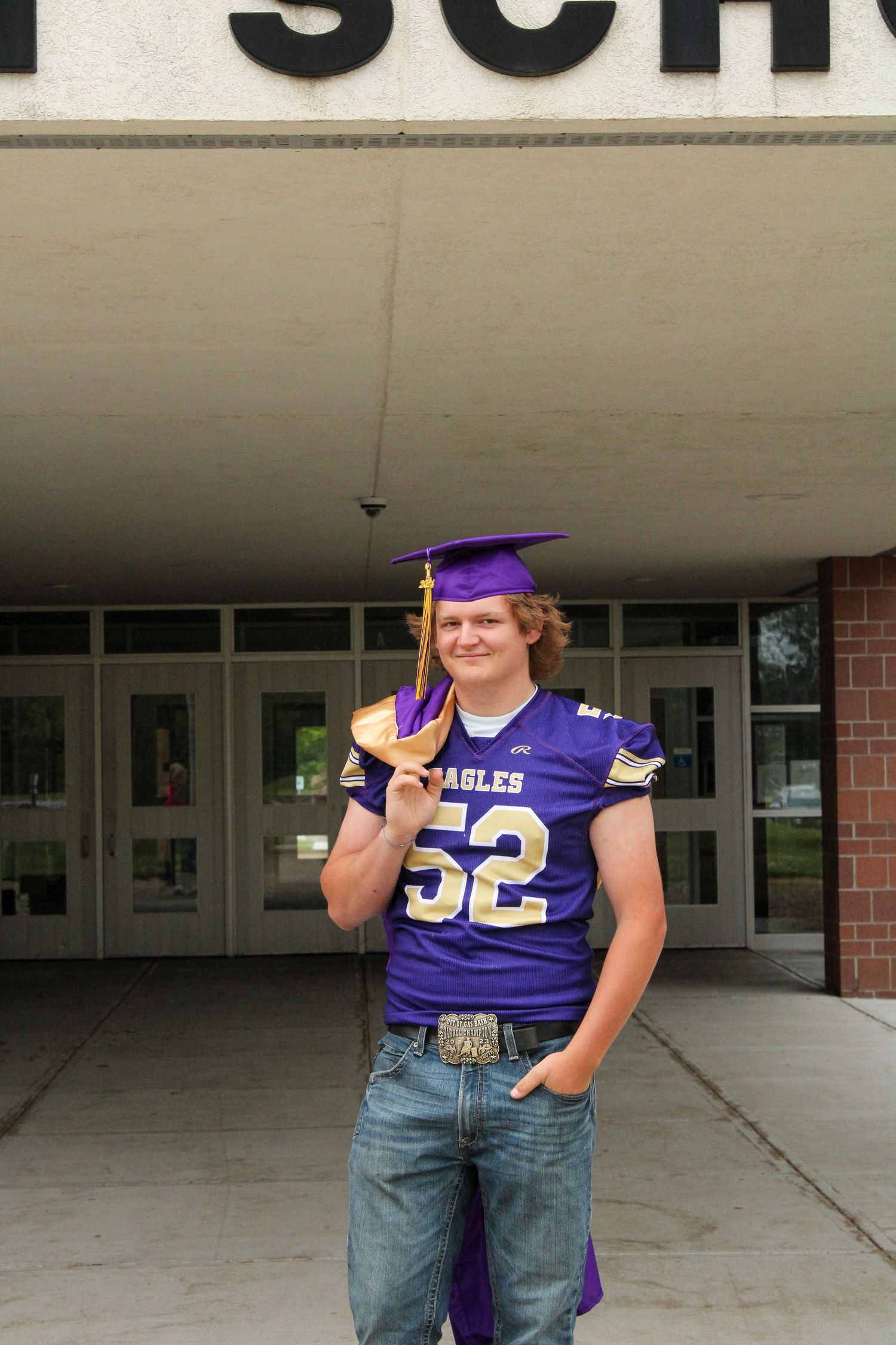 Graduate in purple cap and sports jersey number 52 poses outside school building.