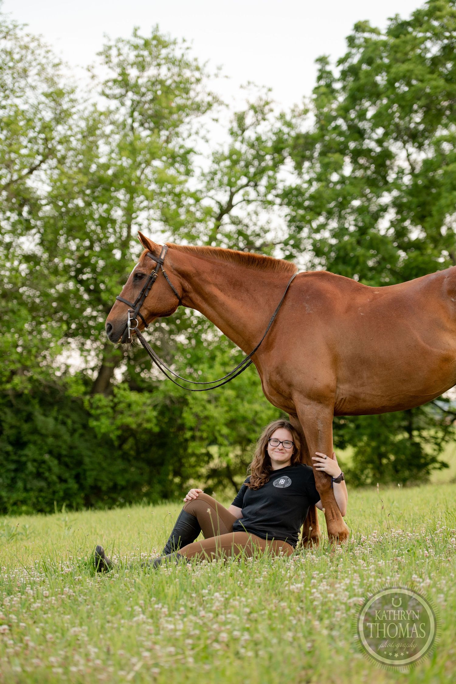 A chestnut horse stands beside someone relaxing in a grassy field with trees in the background.