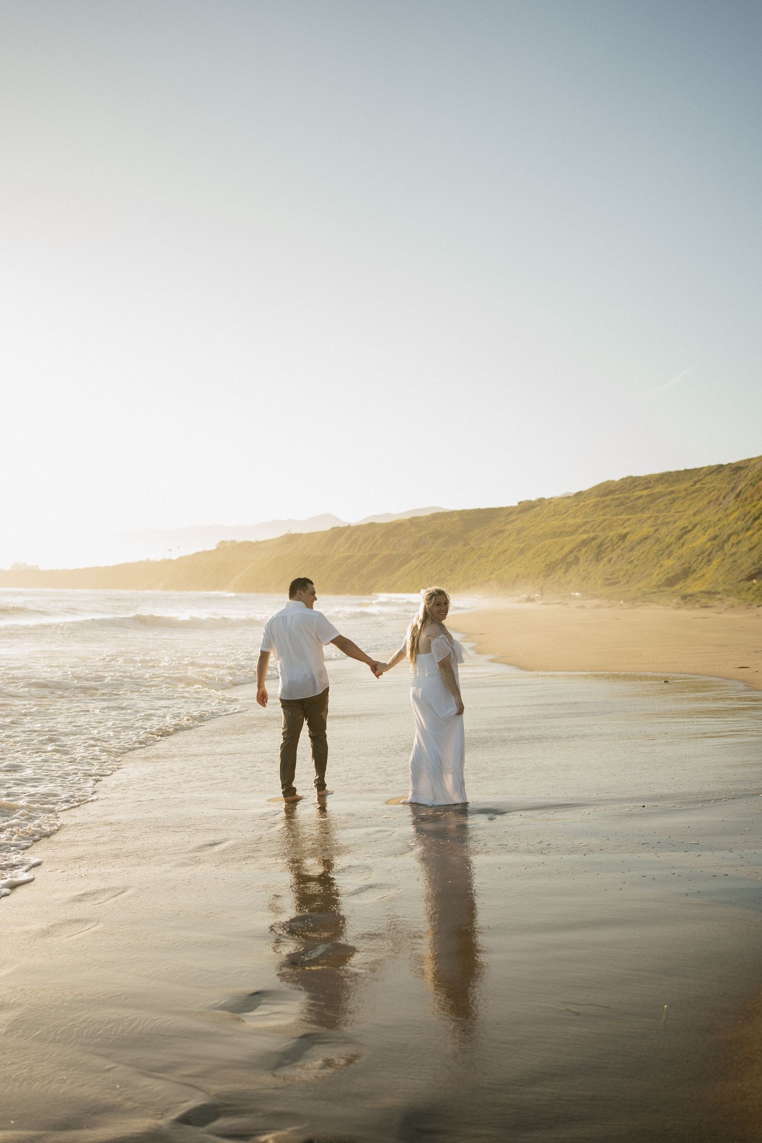 A romantic couple holds hands while walking along a sunset beach with coastal cliffs in the background.