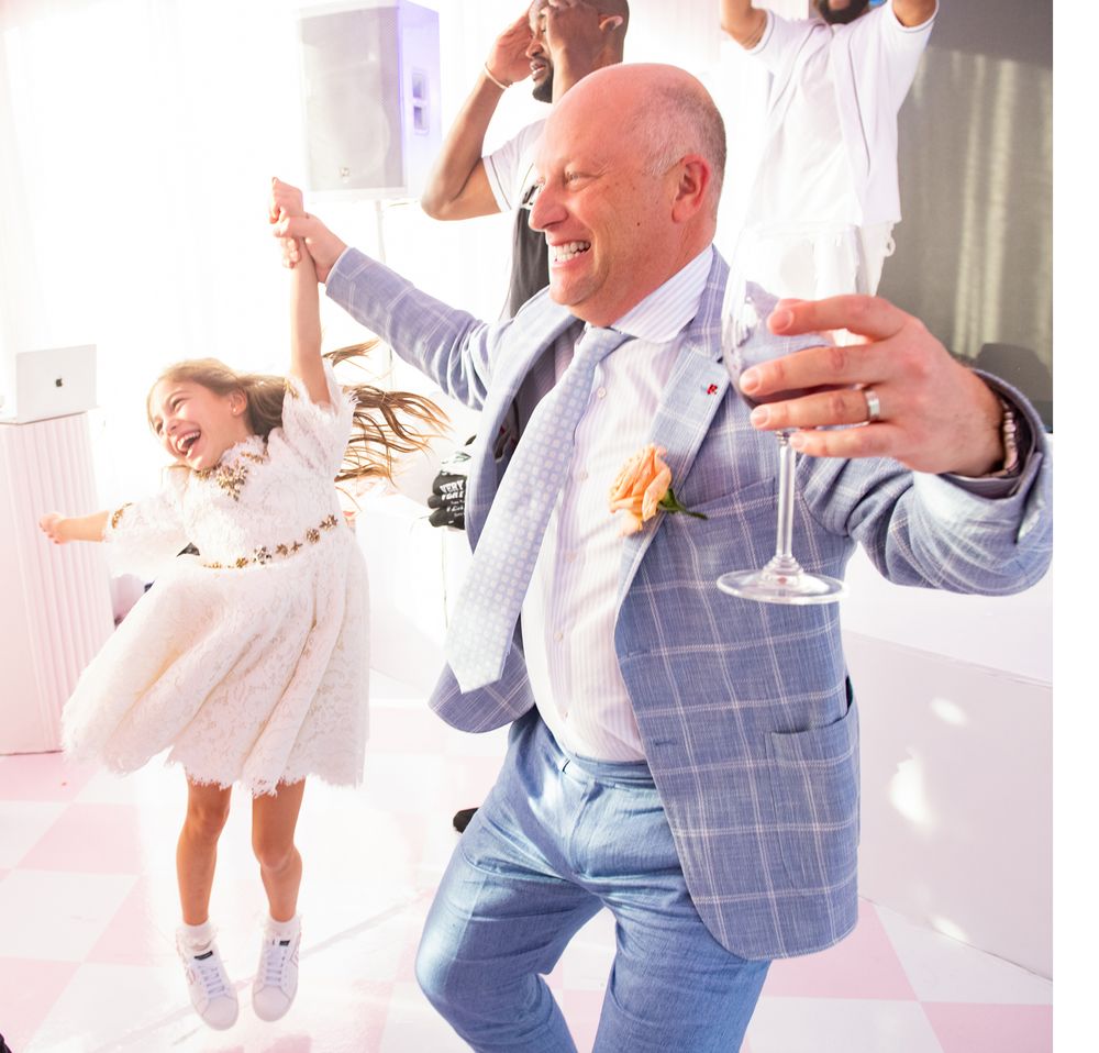 Guests dancing joyfully at an event with pink lighting and white decor.