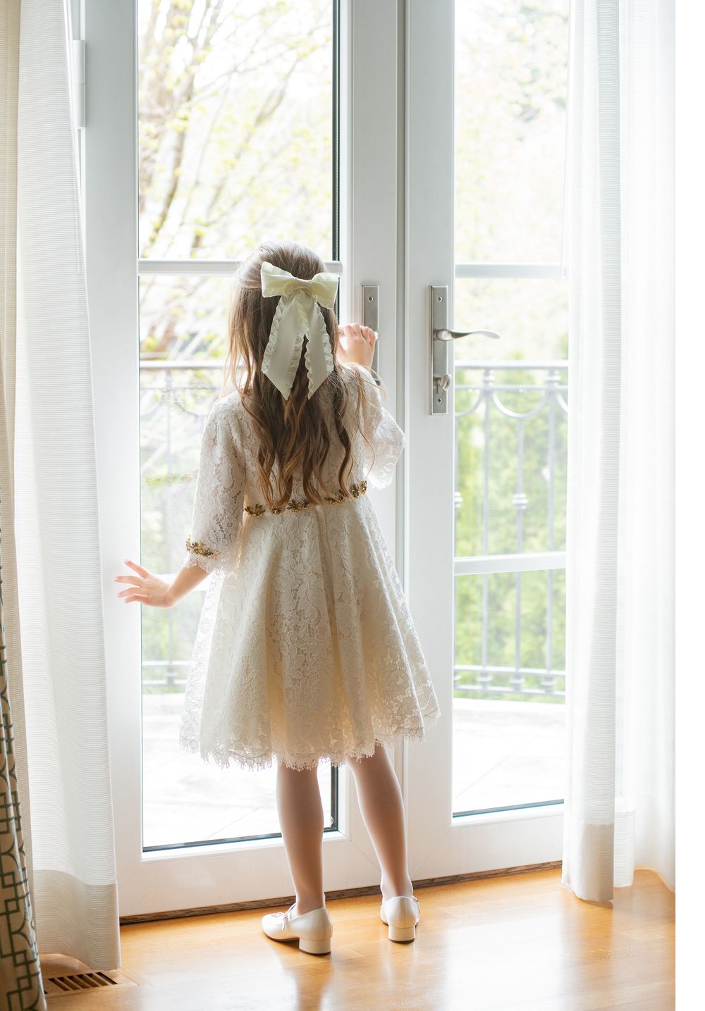 Young dancer in white lace dress looking out bright window with bow in hair.