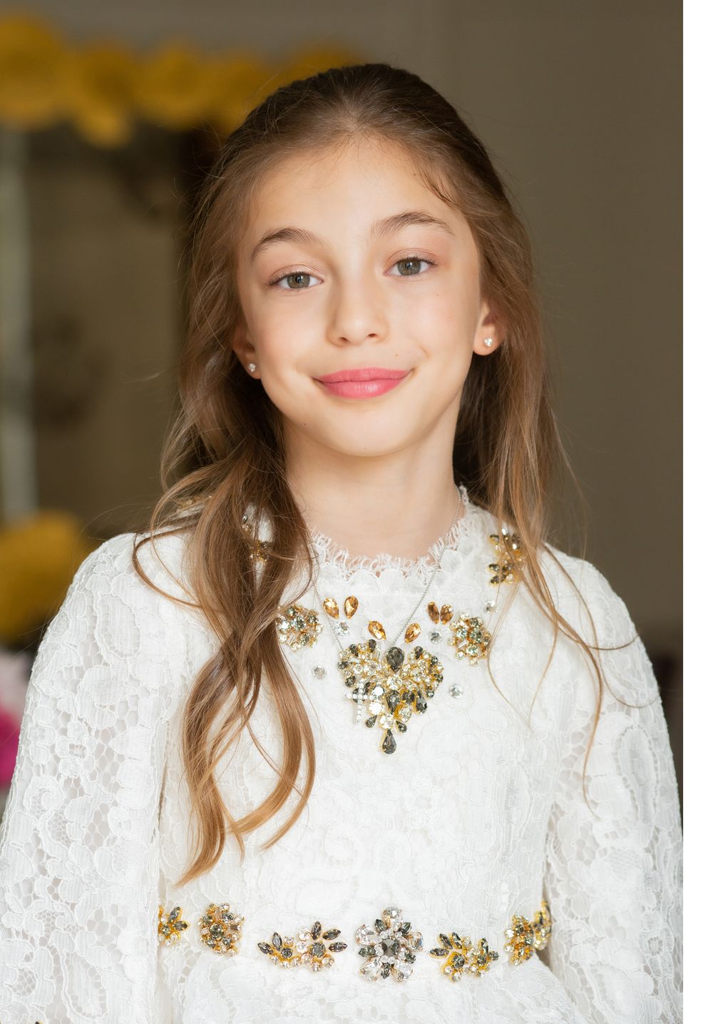Young girl in elegant white lace dress with gold floral embellishments and statement necklace.