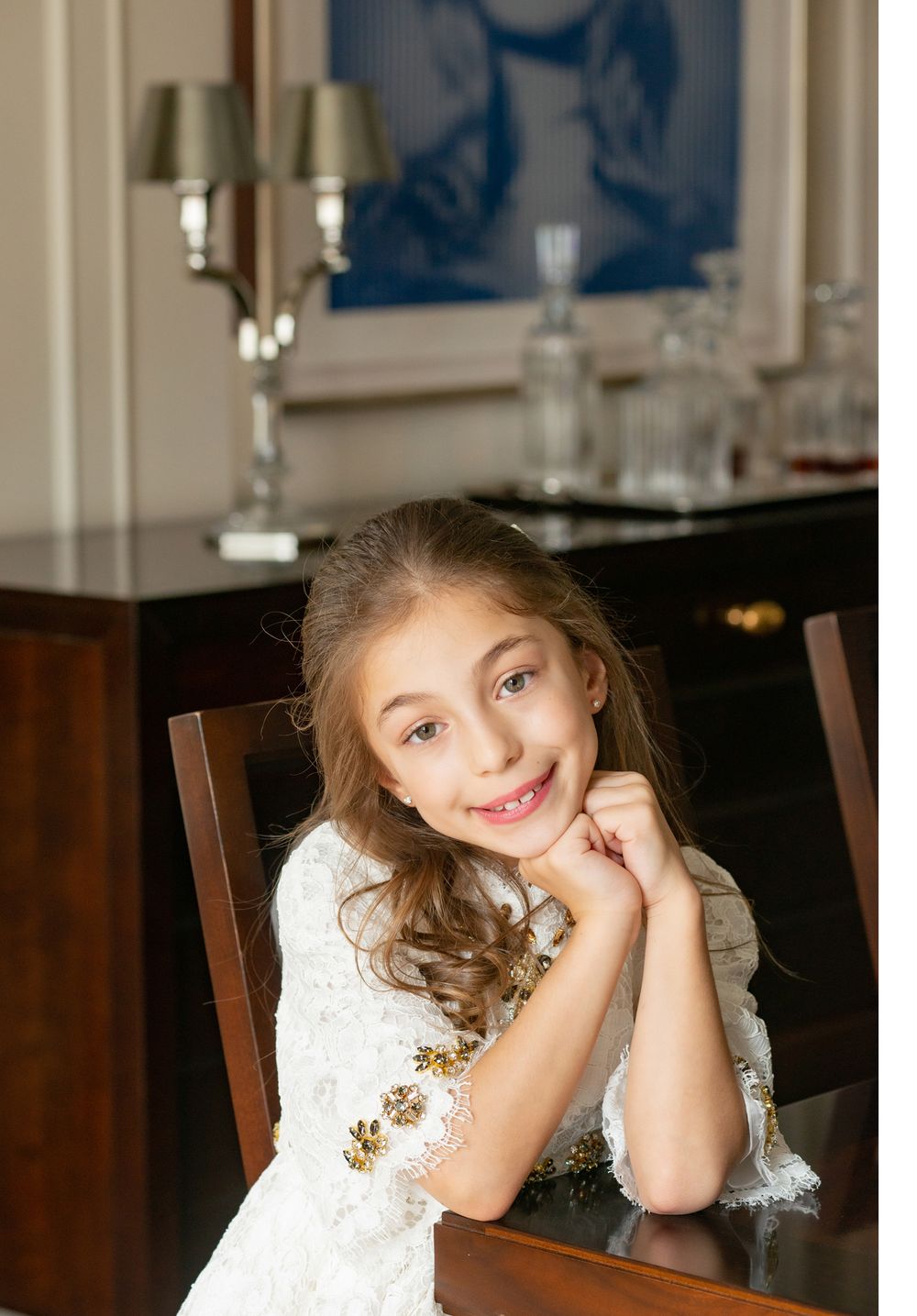 Portrait in front of dining room decor wearing white lace dress with gold accents.