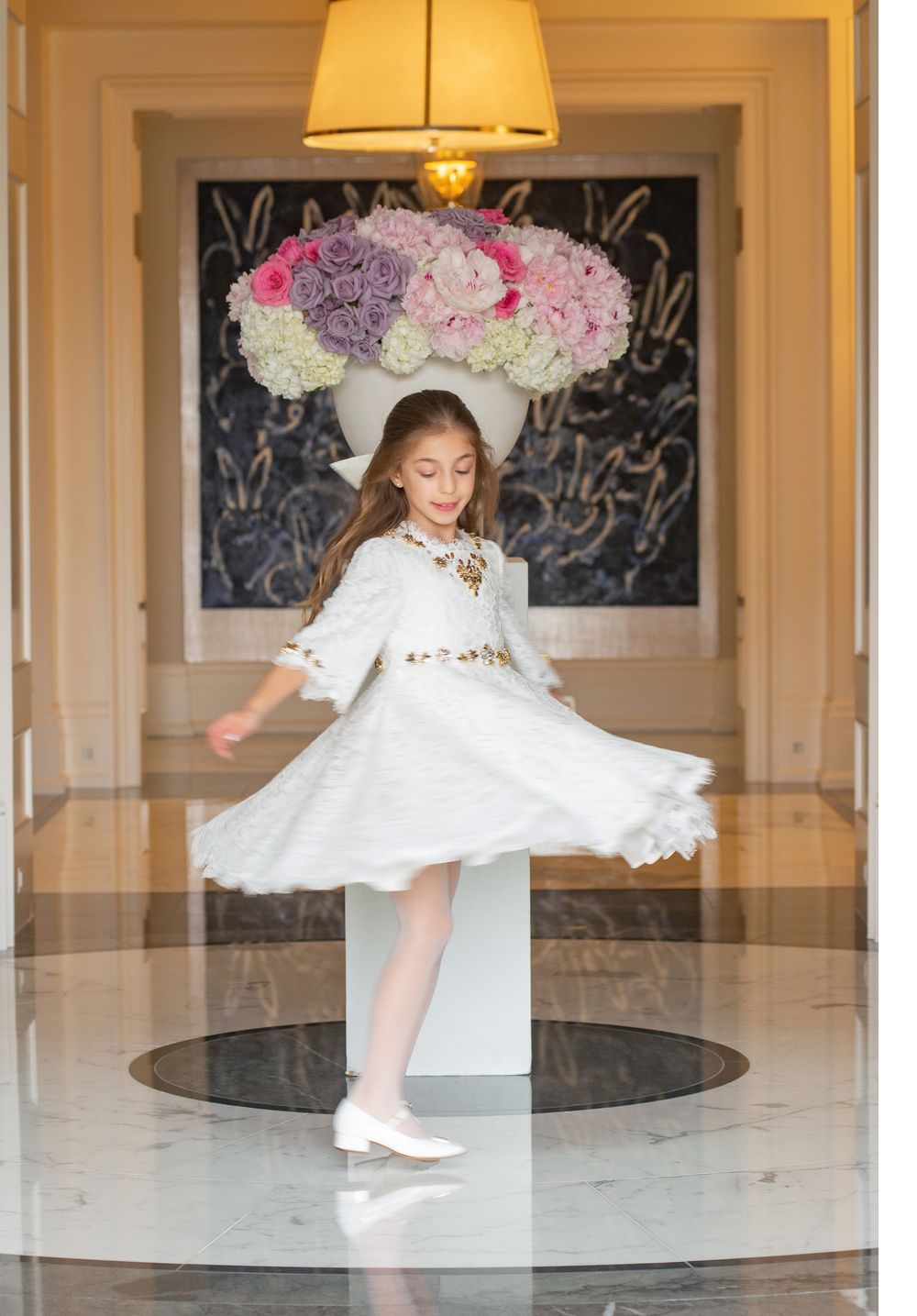 White lace dress twirling beneath floral arrangement and warm lighting in elegant hallway.