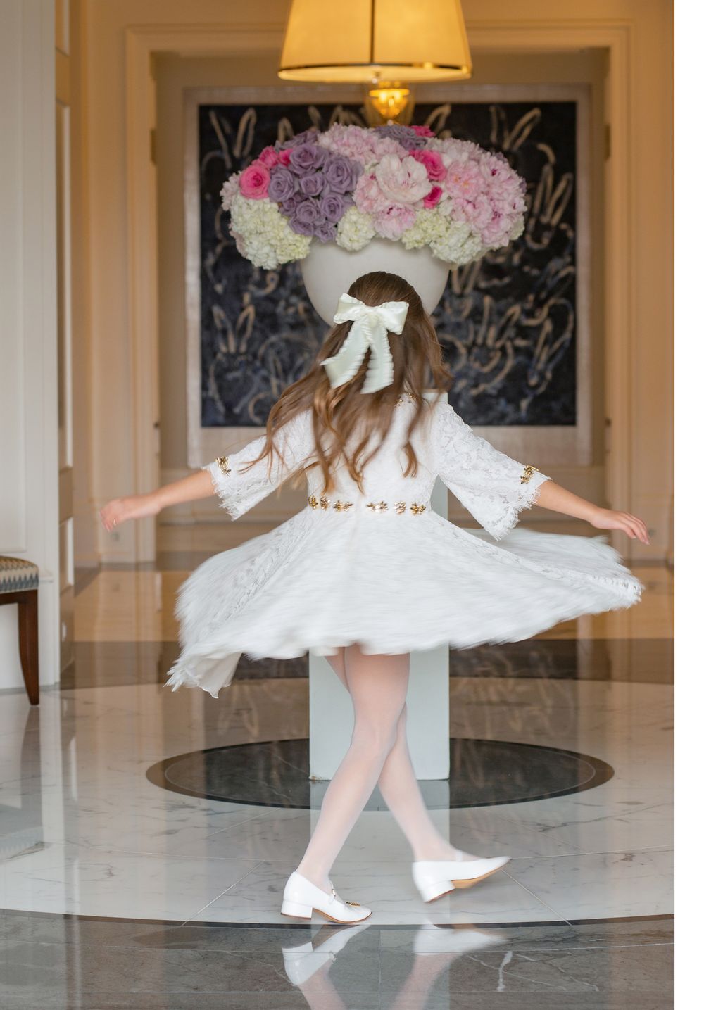 Spinning dance pose in white dress beneath large floral arrangement in elegant interior.