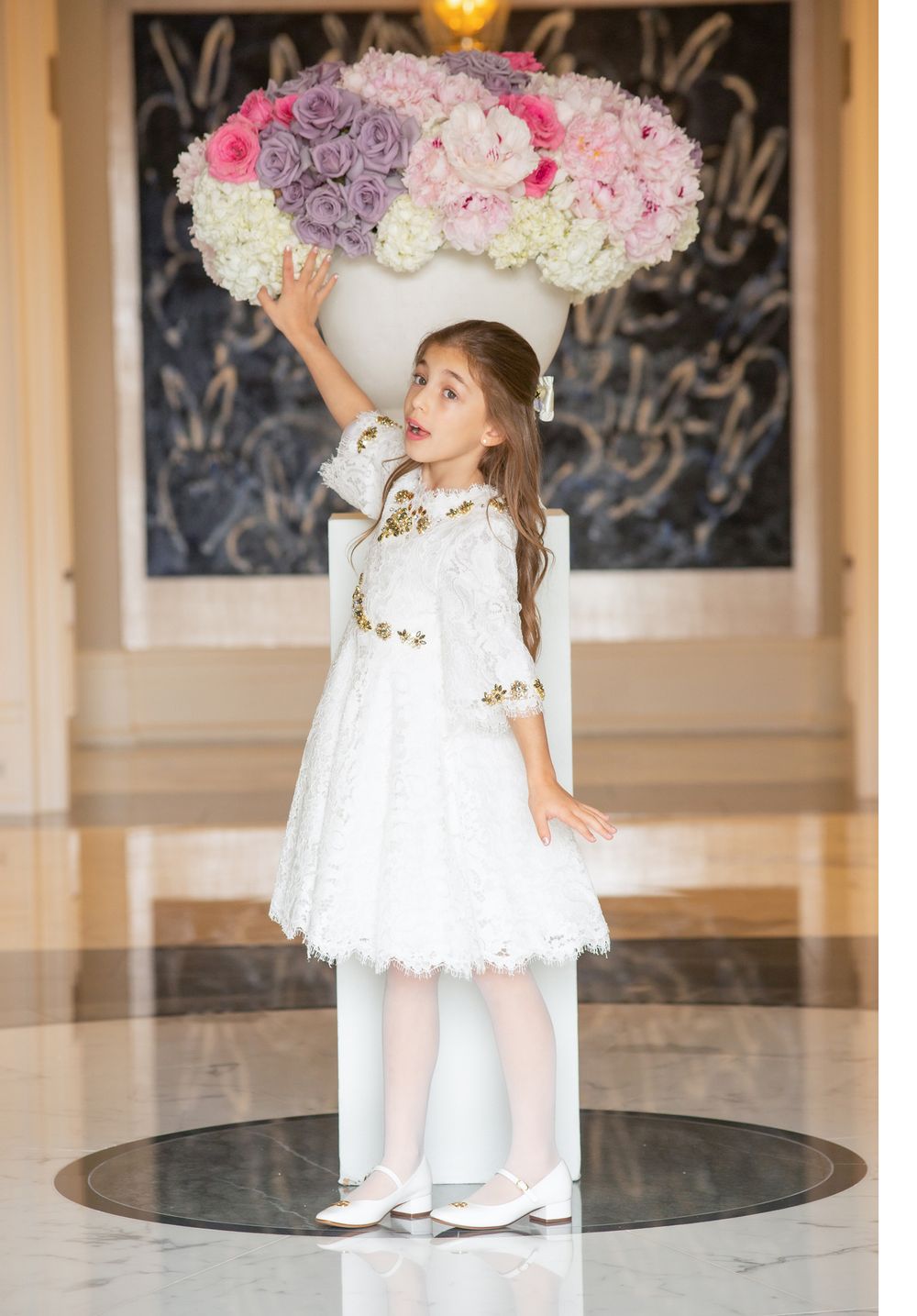 A white lace dress adorned with gold details stands beside an elegant floral arrangement in pink and purple.