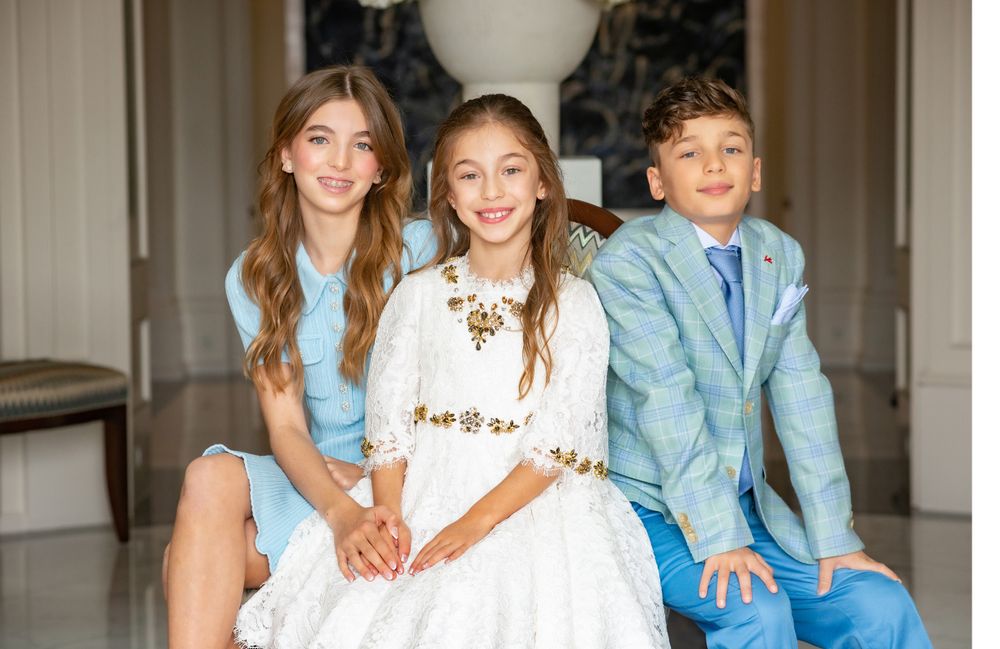 Three children in coordinating pastel outfits pose together in an elegant interior setting.
