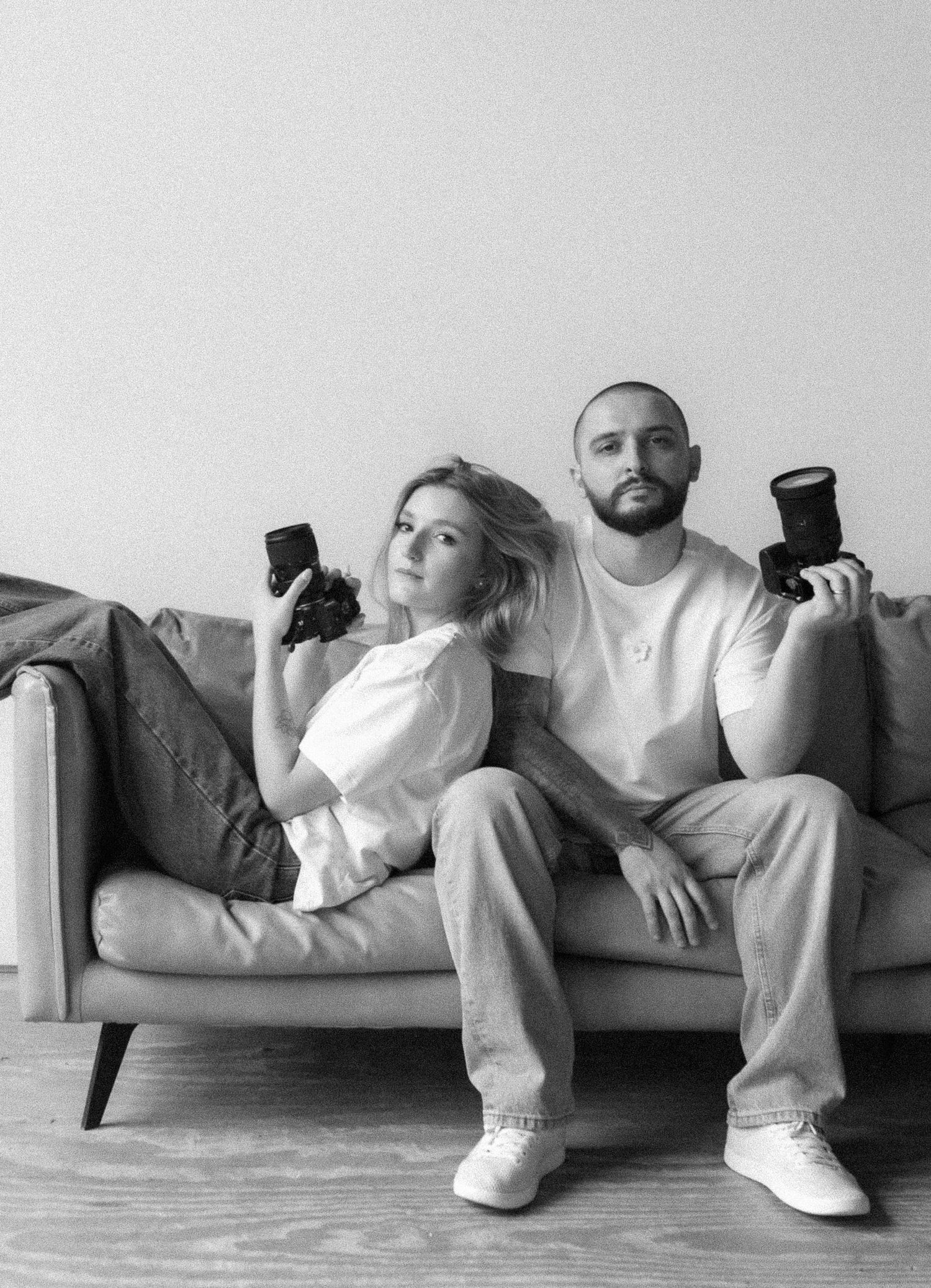 Creative photographers lounging on grey couch with cameras in casual matching outfits.