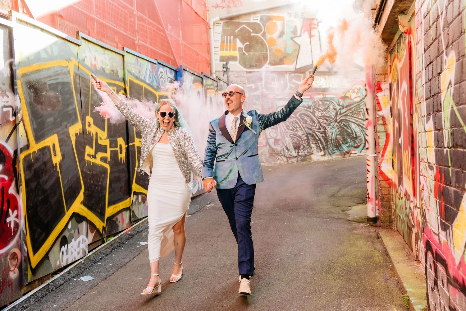 Creative Auckland wedding photo of blue-haired bride & groom in velvet jacket walking through graffiti laneway with coloured smoke bombs