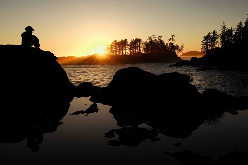 Silhouette of someone sitting on rocks by a lake during sunset with trees in the background.