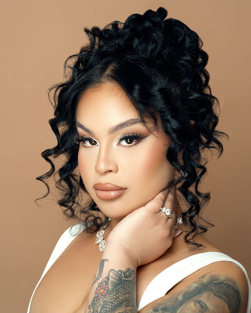 Portrait showcasing glamorous curly updo hairstyle, defined makeup, white top and visible arm tattoos against brown background.