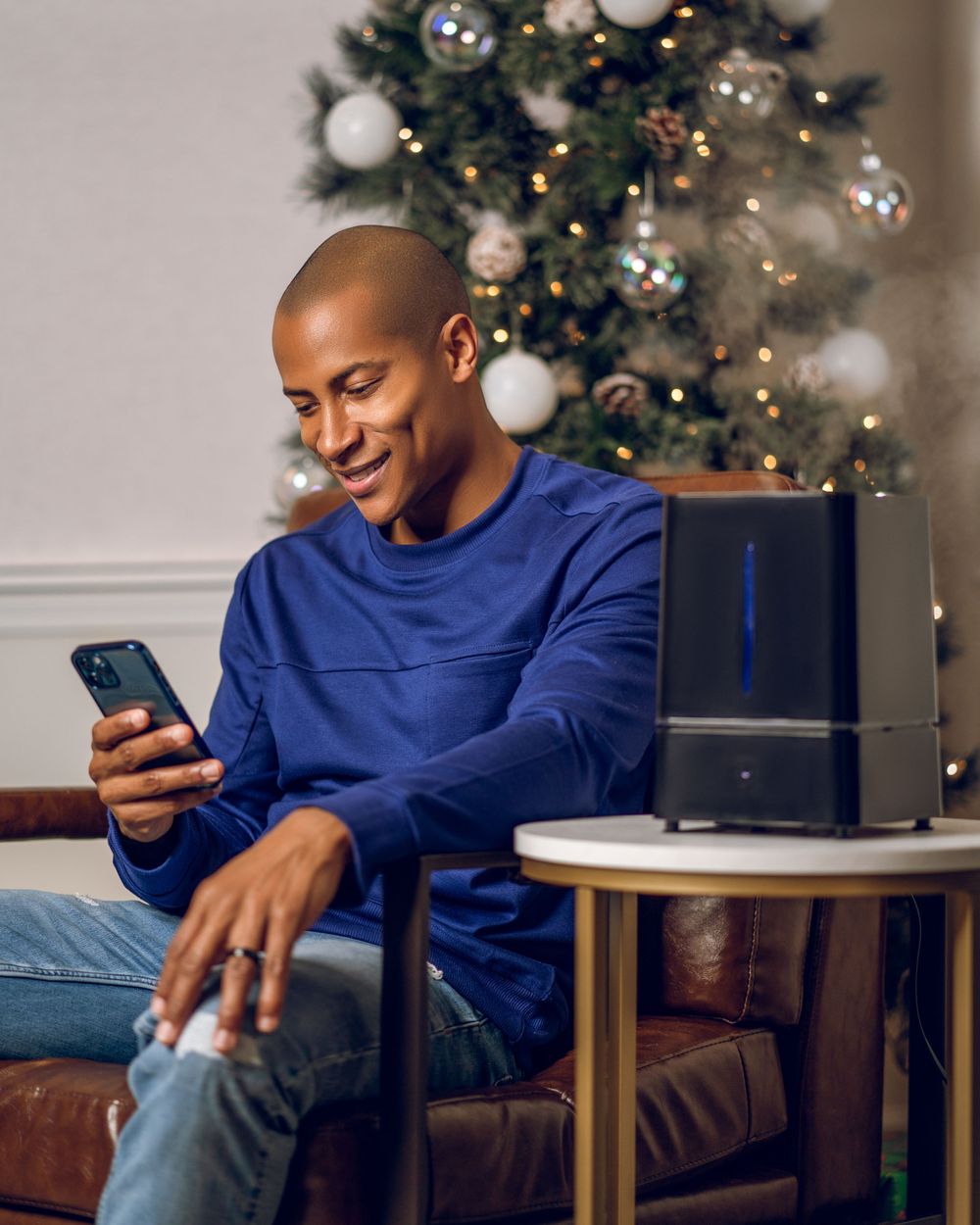 Person relaxes on leather couch while checking phone near decorated Christmas tree.