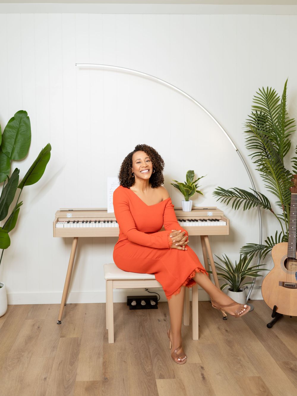 Musician in orange dress sitting at piano surrounded by plants in bright modern studio.
