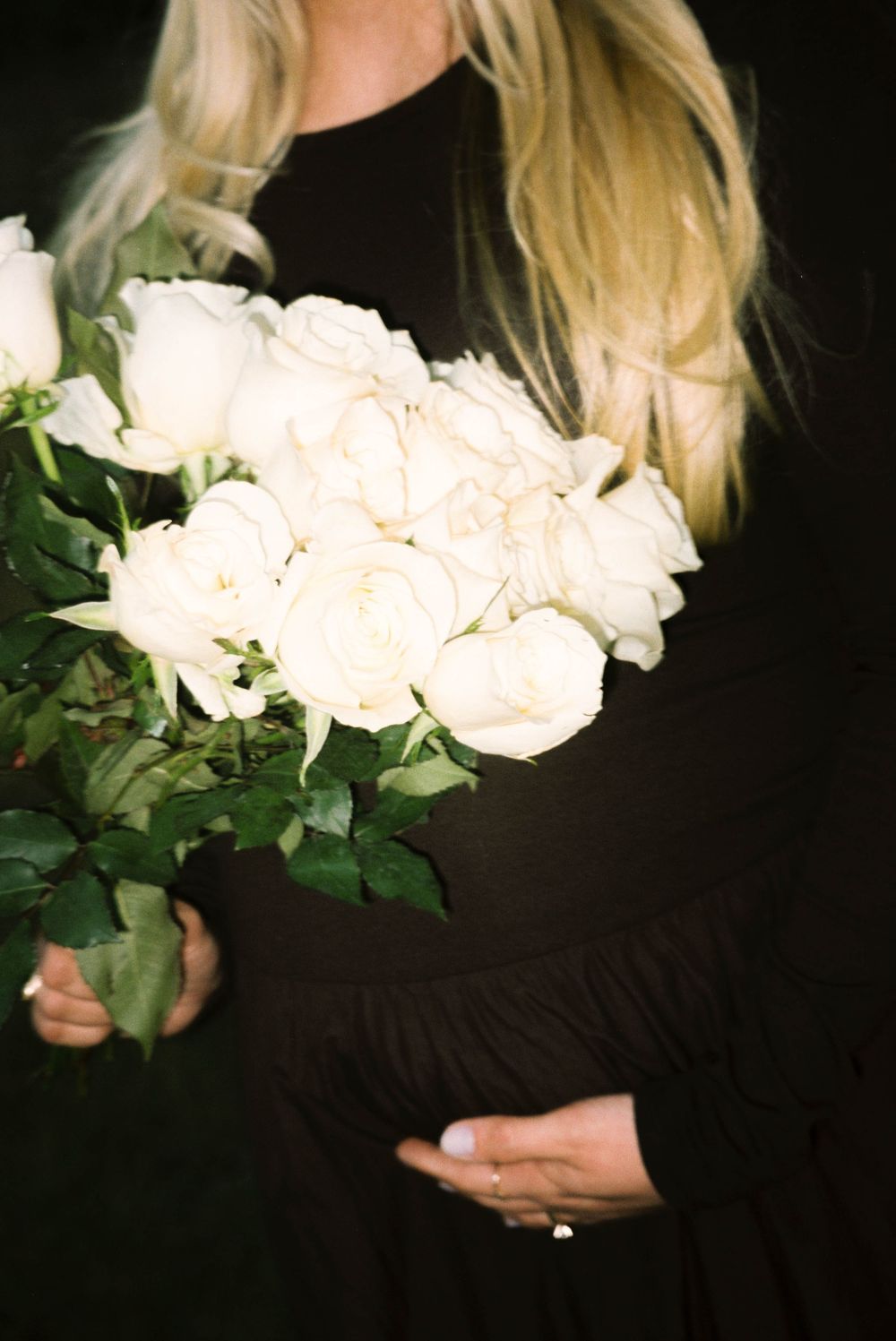 Pregnant woman in black dress holds bouquet of white roses while cradling baby bump.