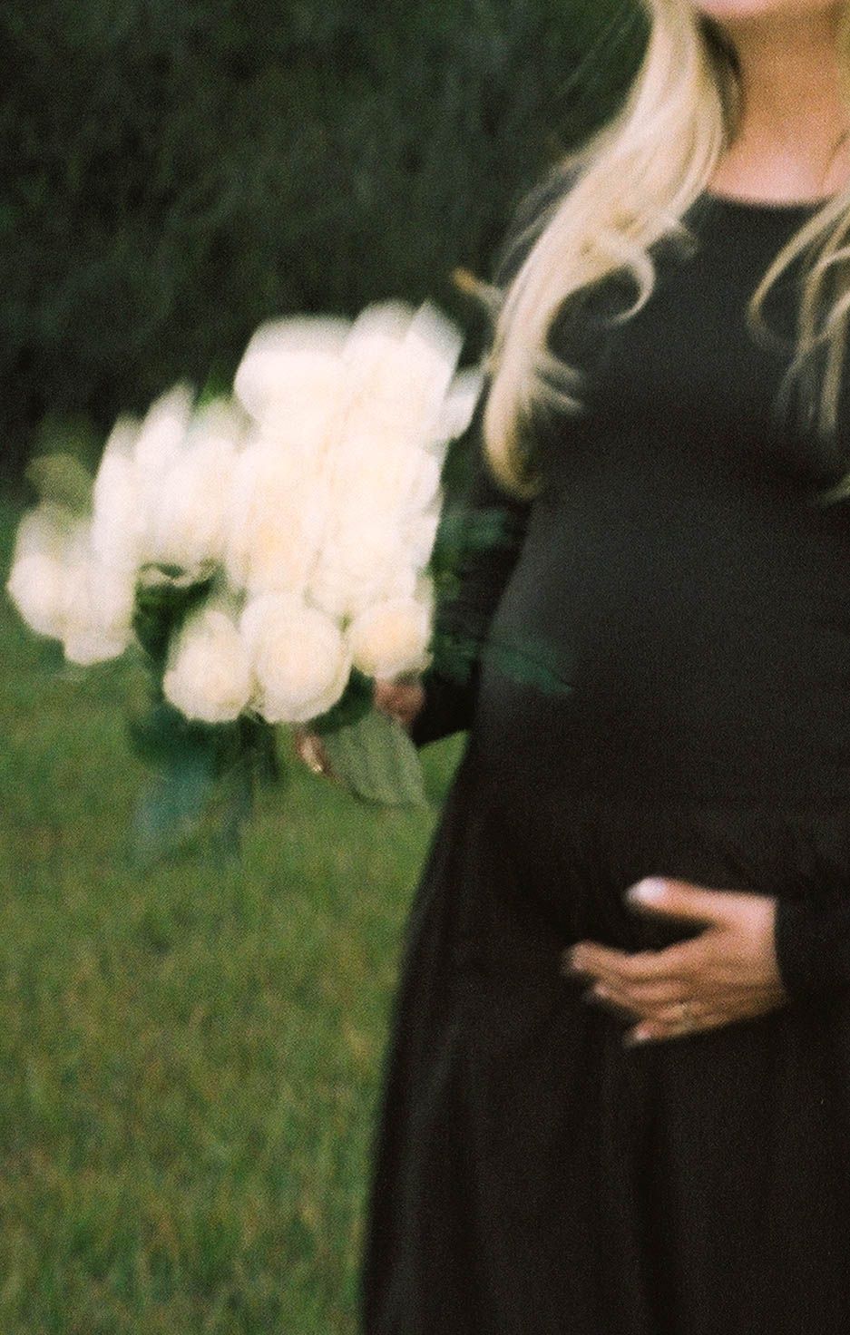 Dreamy motion-blur photo of pregnant woman in black dress holding white roses outdoors.