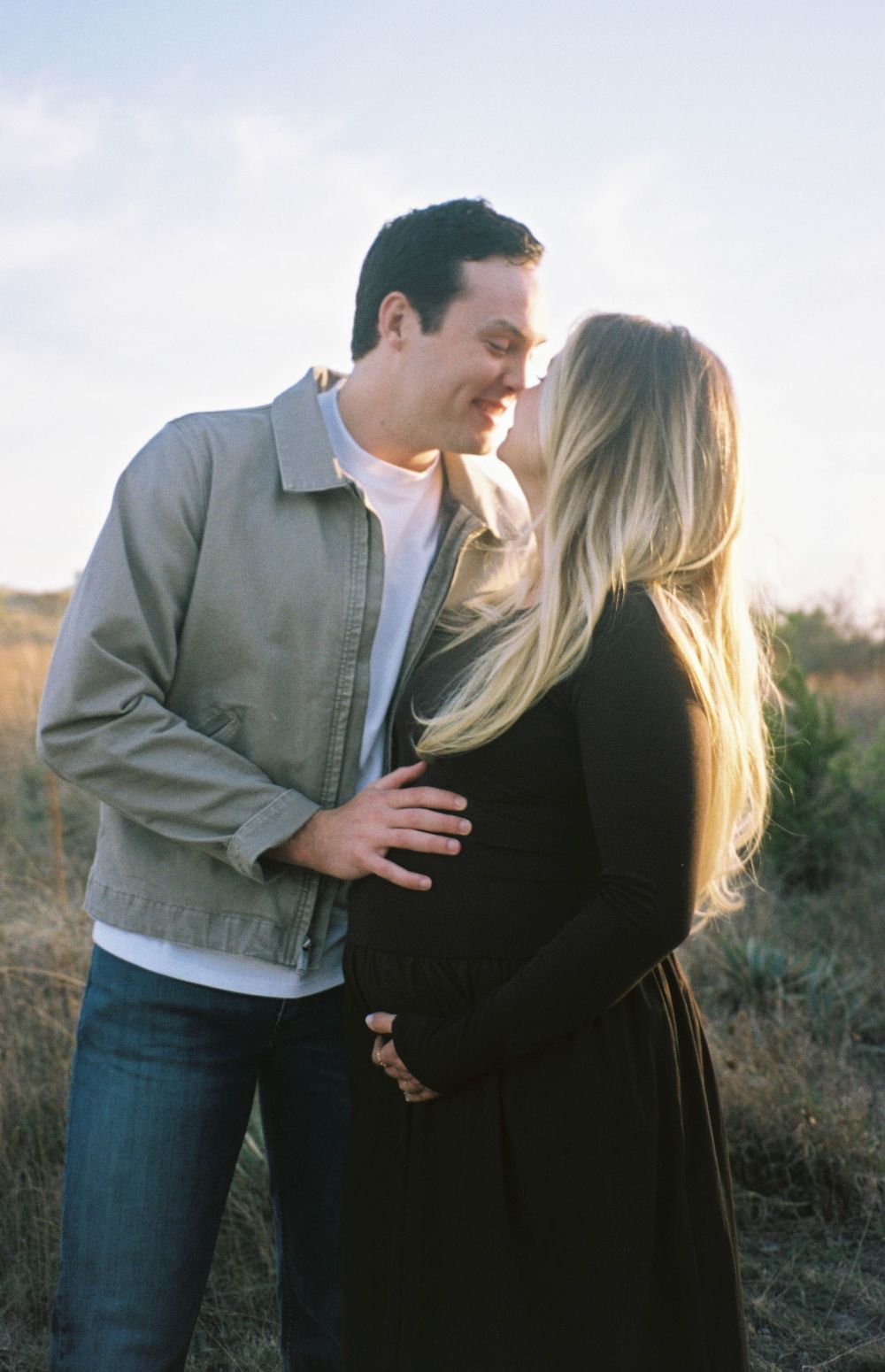 Couple shares tender moment in sunlit field, man gently holding pregnant woman's baby bump.
