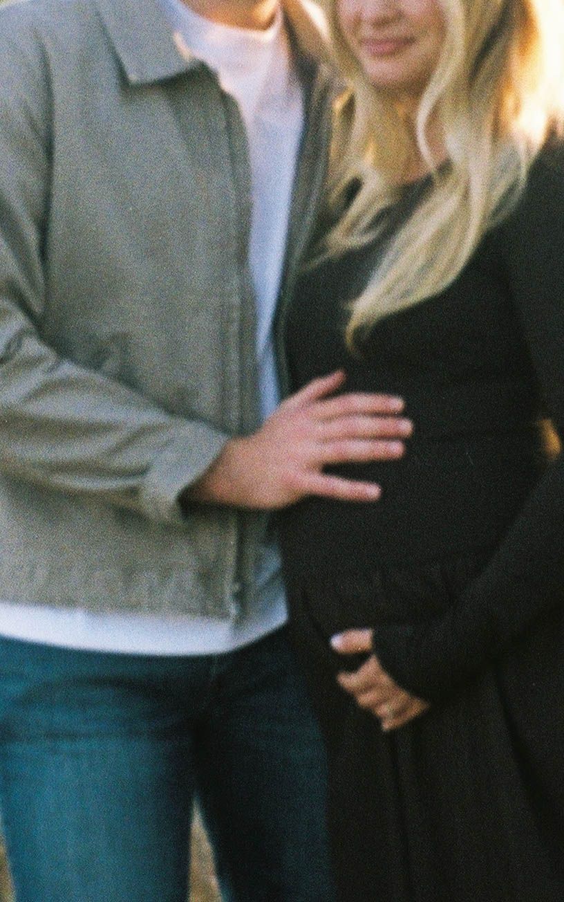 Couple maternity photo with man placing hand on pregnant woman's baby bump outdoors.