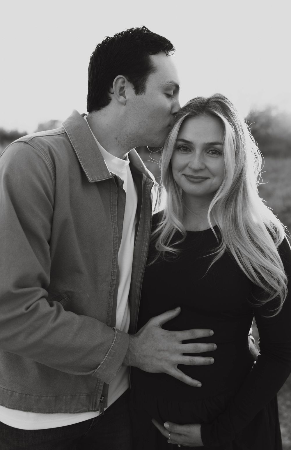 Black and white photo of man kissing pregnant woman's cheek in a field.
