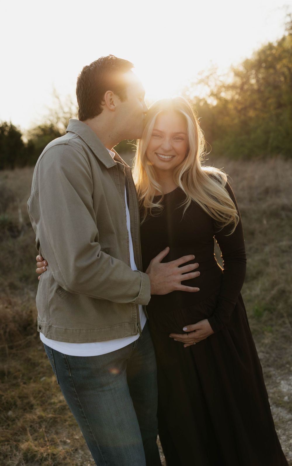 Glowing pregnant couple embracing at golden hour in a sunlit outdoor field.