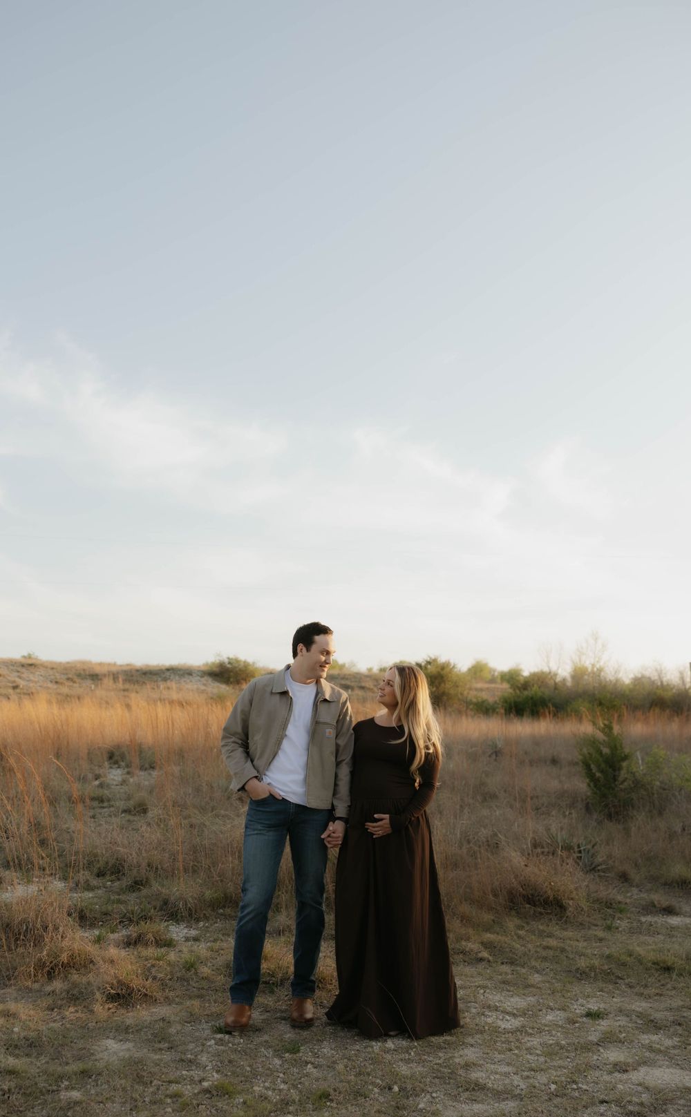 Expecting couple holding hands in an open field under a soft cloudy sky.