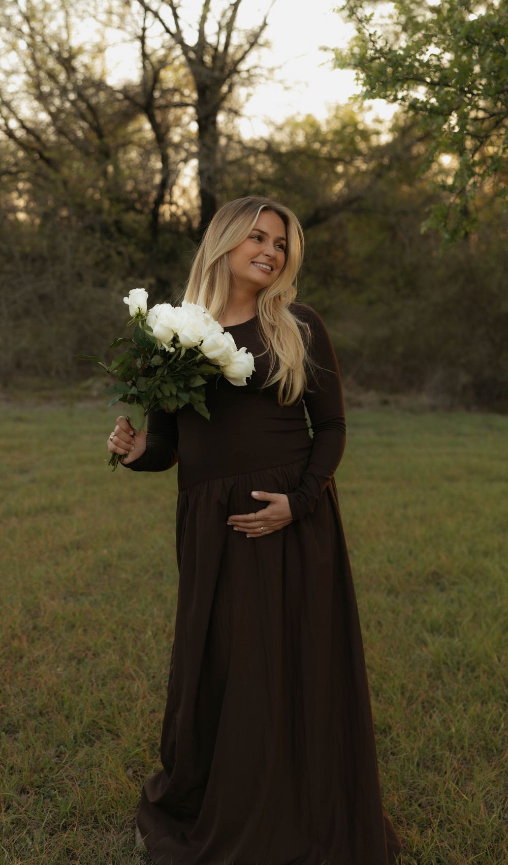 Radiant pregnant woman in brown maxi dress holding white flowers, smiling in a sunlit park.