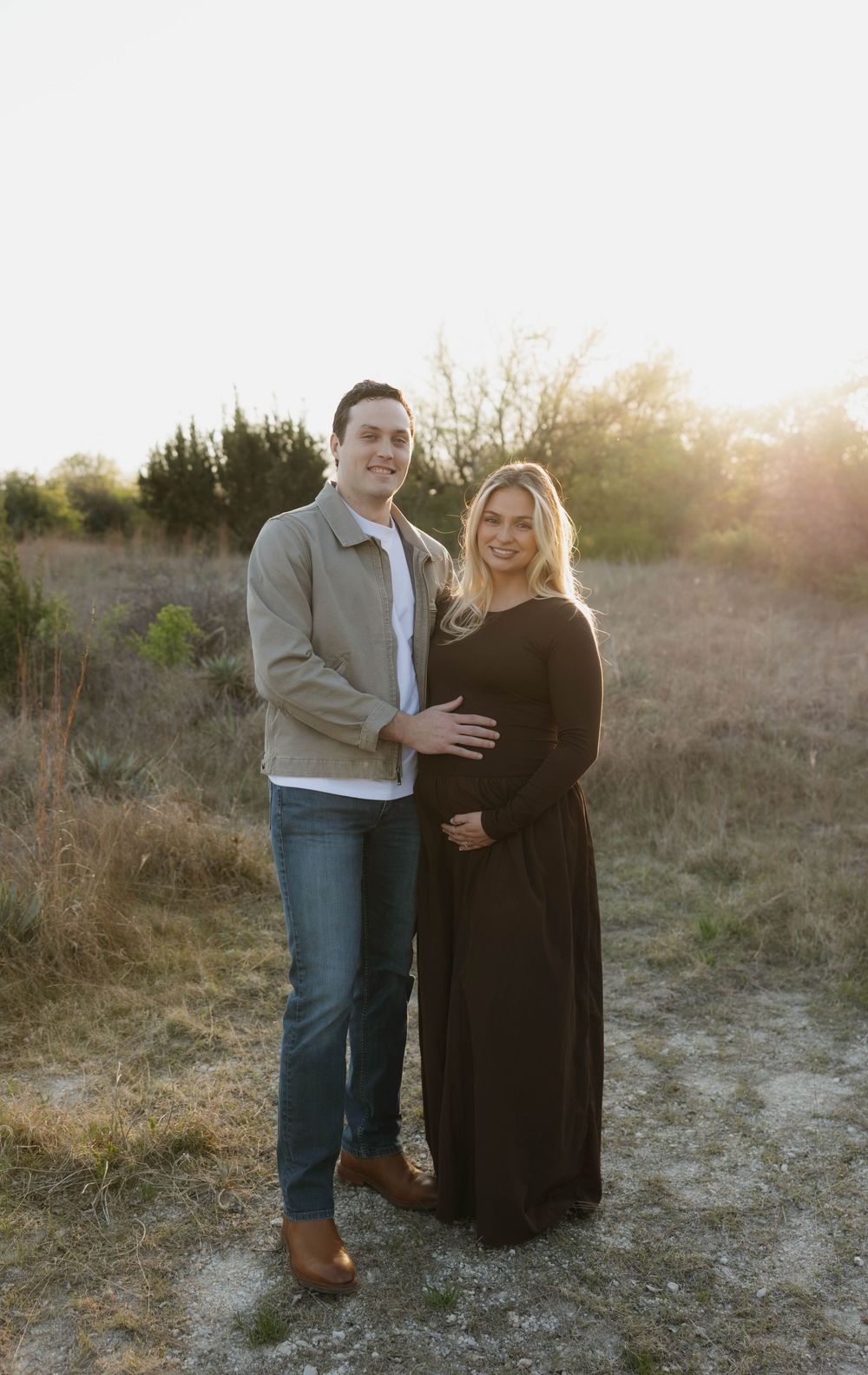 Smiling couple poses together in golden hour field, woman in black maternity gown showing baby bump.