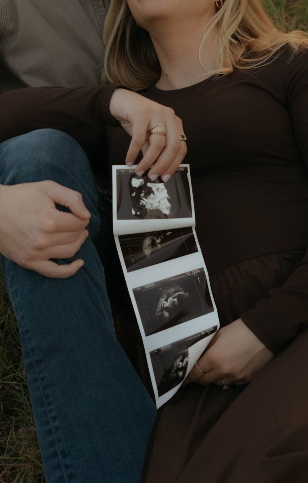 Pregnant woman holding ultrasound photos while sitting outdoors with partner during maternity photo session.