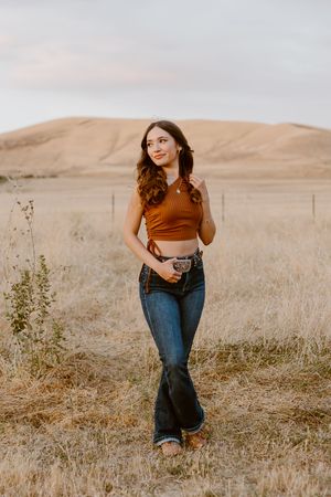 Young woman in rust crop top and flare jeans poses in golden field with rolling hills at sunset.