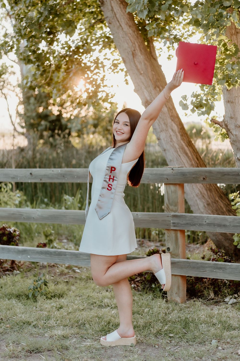 Young woman in white dress and graduation sash holds red cap aloft outdoors, celebrating high school graduation.