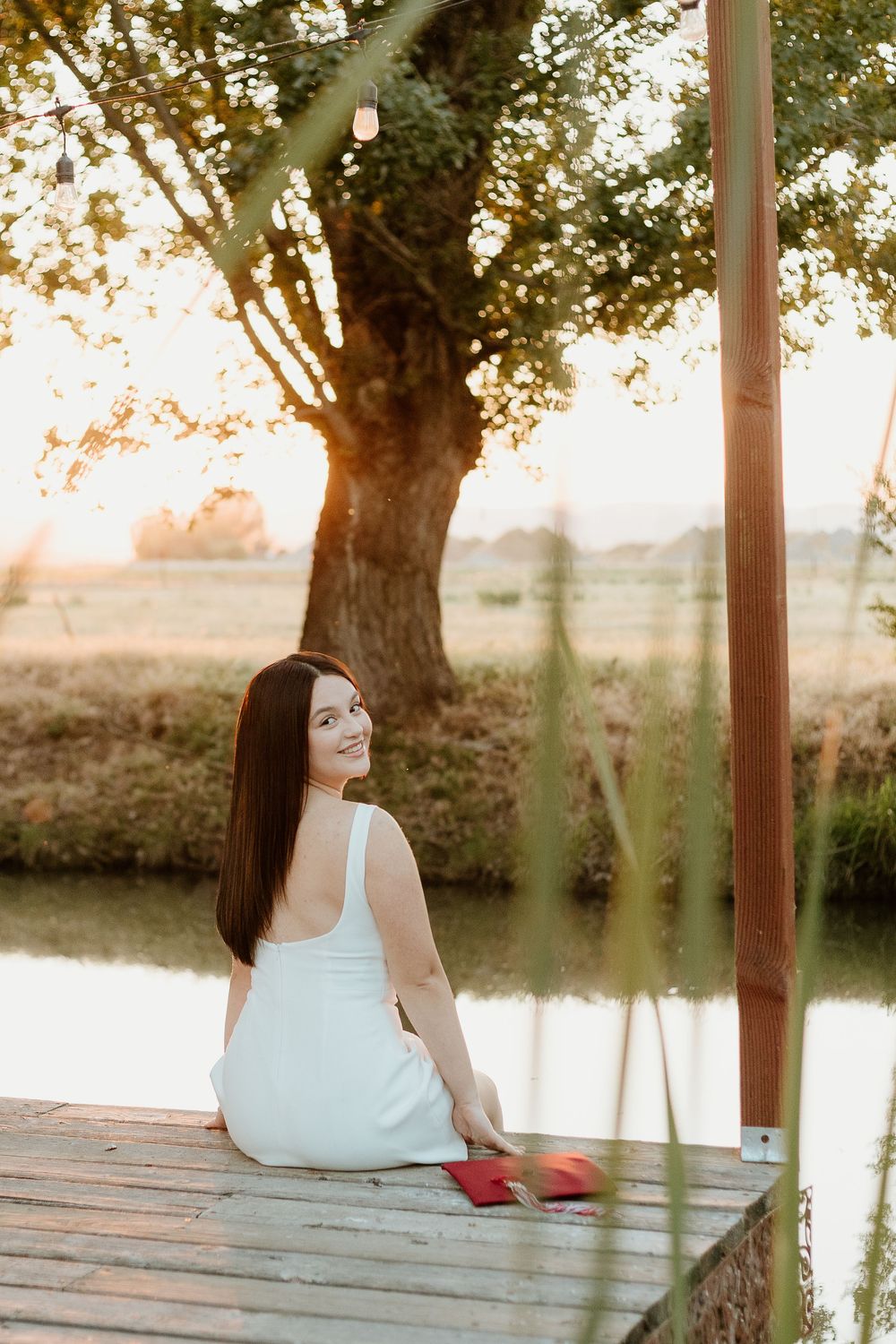 Young woman in white dress sitting on wooden dock by calm lake at golden sunset, smiling over shoulder.