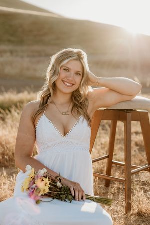 Smiling blonde woman in white dress holding colorful bouquet, seated outdoors in golden sunlight with rolling hills backdrop.