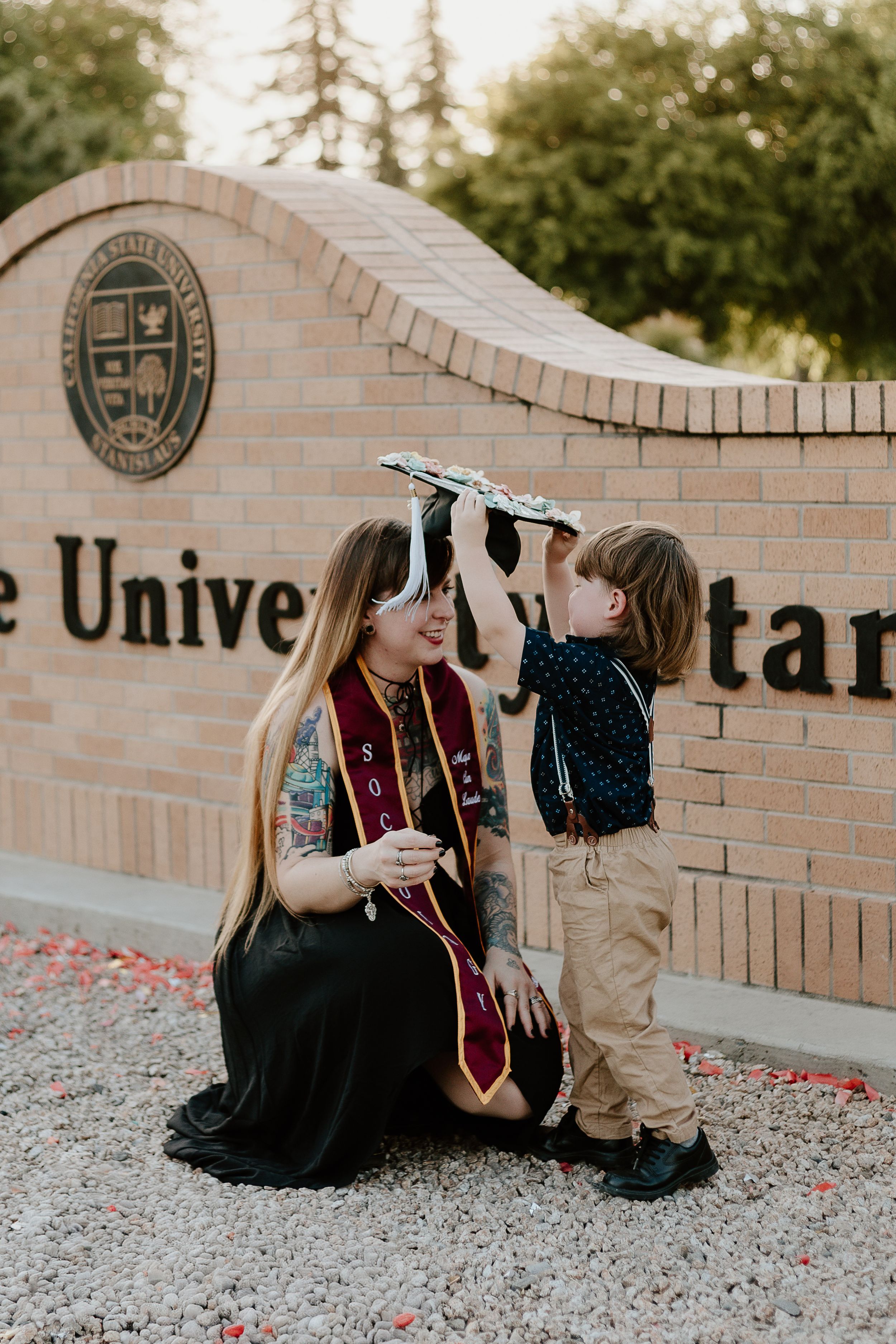 A graduate in cap and gown kneels as a young boy places her graduation cap on her head in front of a university sign.