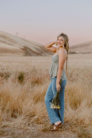 Young blonde woman in green top and jeans holding wildflowers in a golden field at sunset.