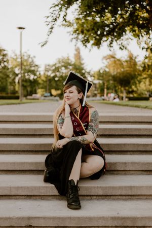 A young woman in graduation cap and gown sits on stone steps, showcasing tattoos, smiling thoughtfully outdoors.