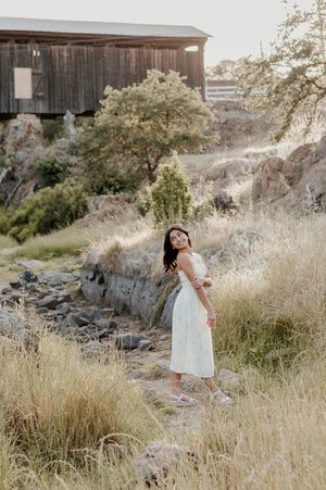 Woman in white midi dress standing among rocks and tall grass near rustic wooden barn in golden natural light.