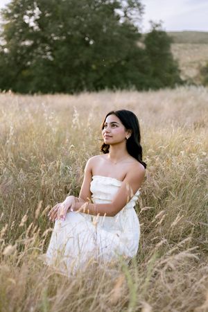 Young woman in white strapless dress sitting gracefully in golden field at sunset.