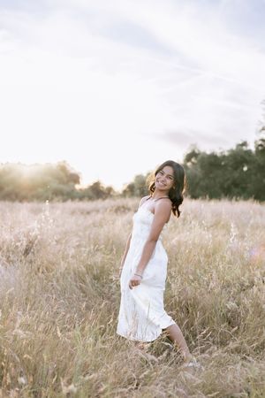 Young woman in white sundress walking barefoot through golden field at sunset, smiling over shoulder.