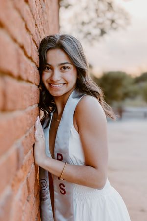 Young woman in white graduation dress leaning against brick wall, smiling outdoors at golden hour.