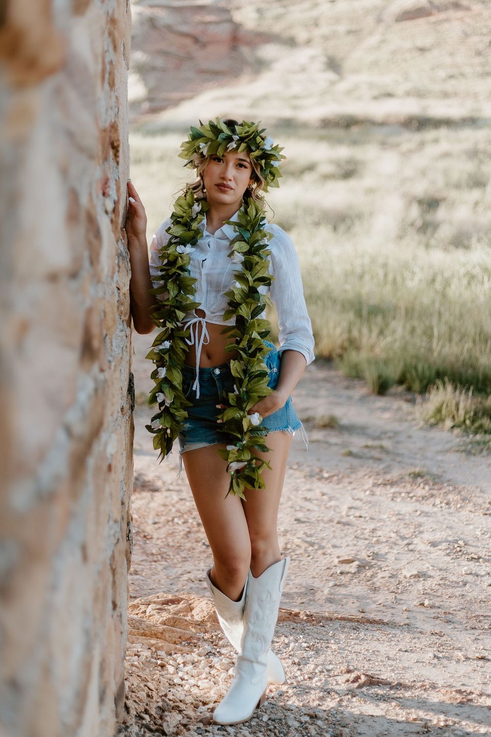Woman in denim shorts, white crop top, cowboy boots, and floral lei and crown poses against a rustic stone wall outdoors.
