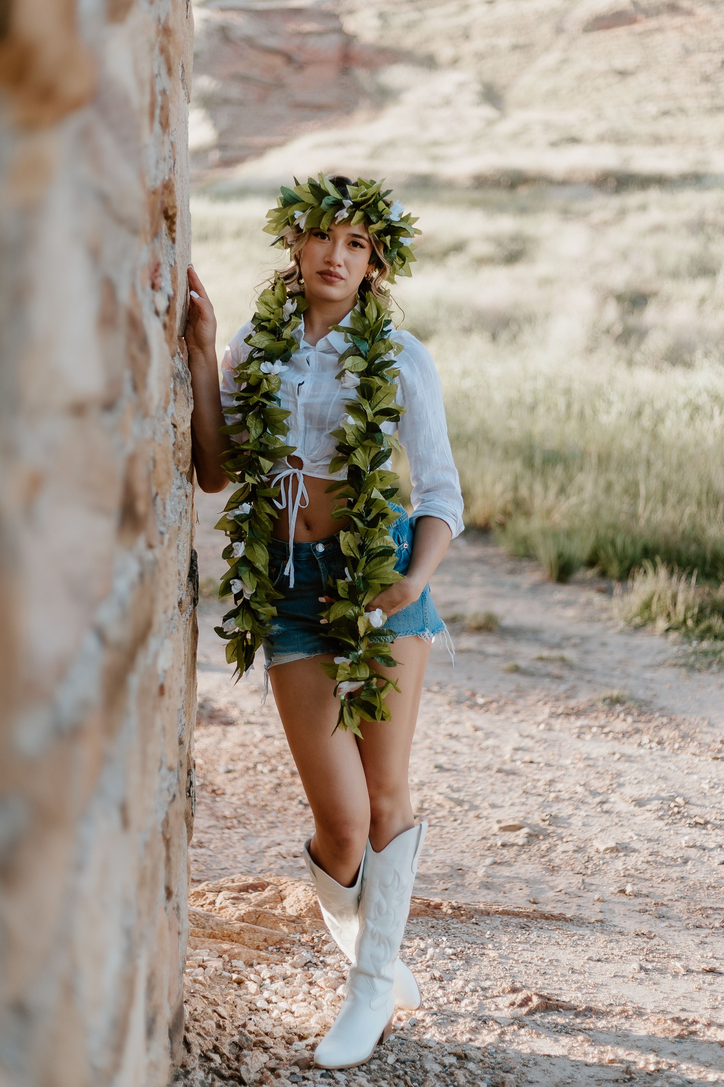 Woman in denim shorts, white crop top, cowboy boots, and floral lei and crown poses against a rustic stone wall outdoors.
