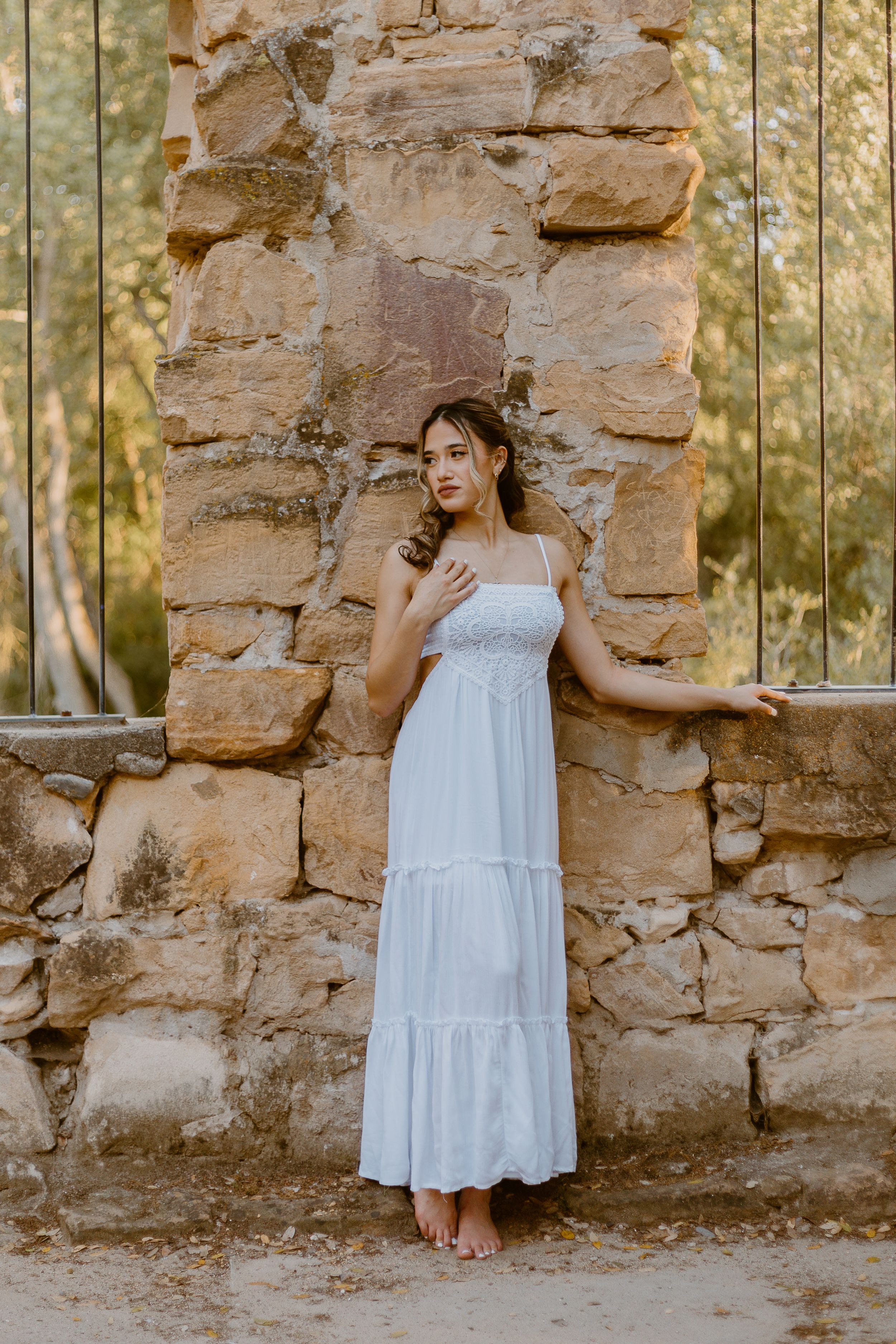 A young woman in a flowing white maxi dress poses gracefully against a rustic stone wall outdoors.