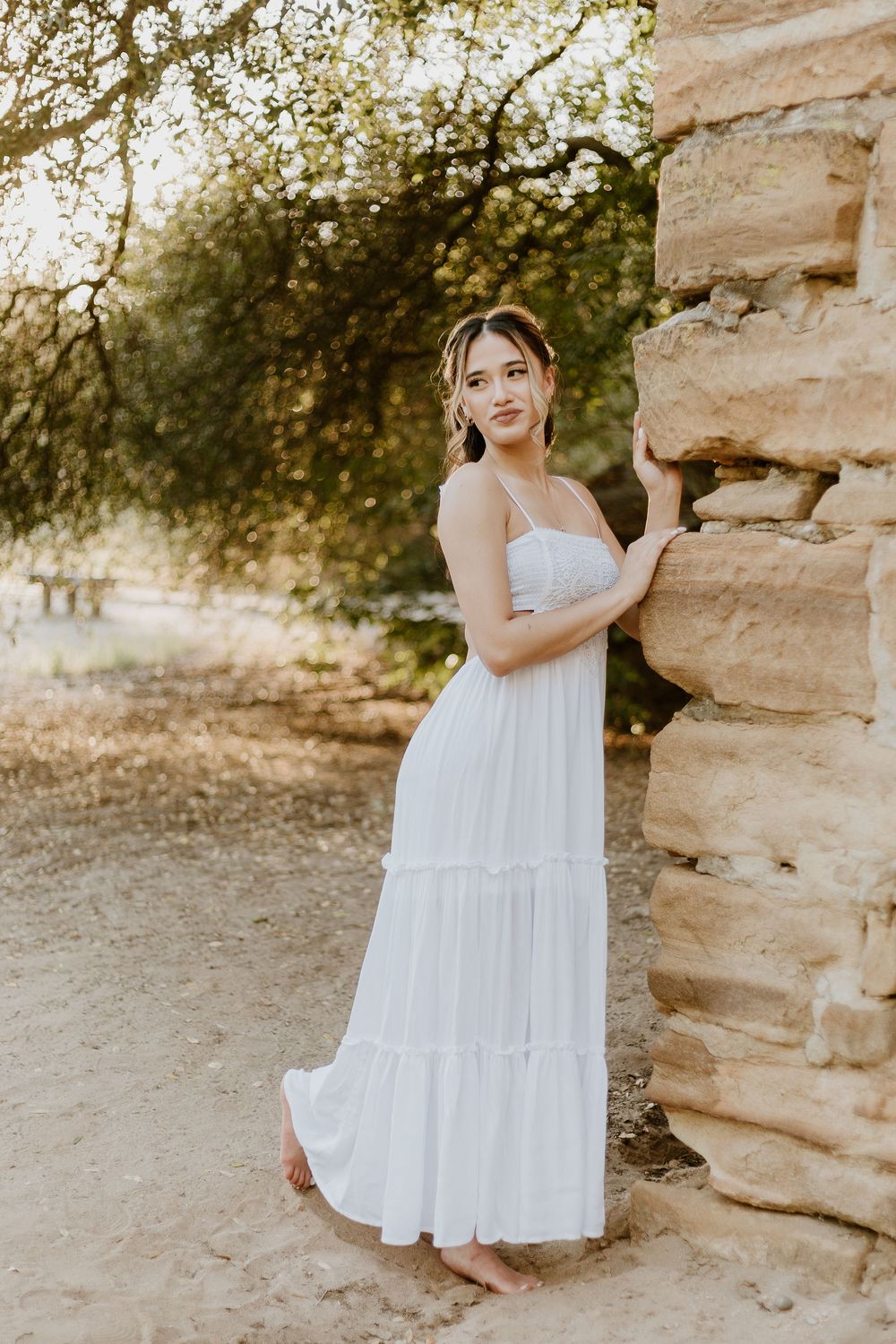 Young woman in white maxi dress poses barefoot beside rustic stone wall surrounded by trees.