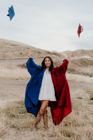 Smiling graduate wearing blue and red robes tosses caps in the air outdoors in a scenic hilly landscape.