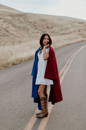 Young woman in white dress, blue and red cape, cowboy boots, standing on rural road with golden hills backdrop.
