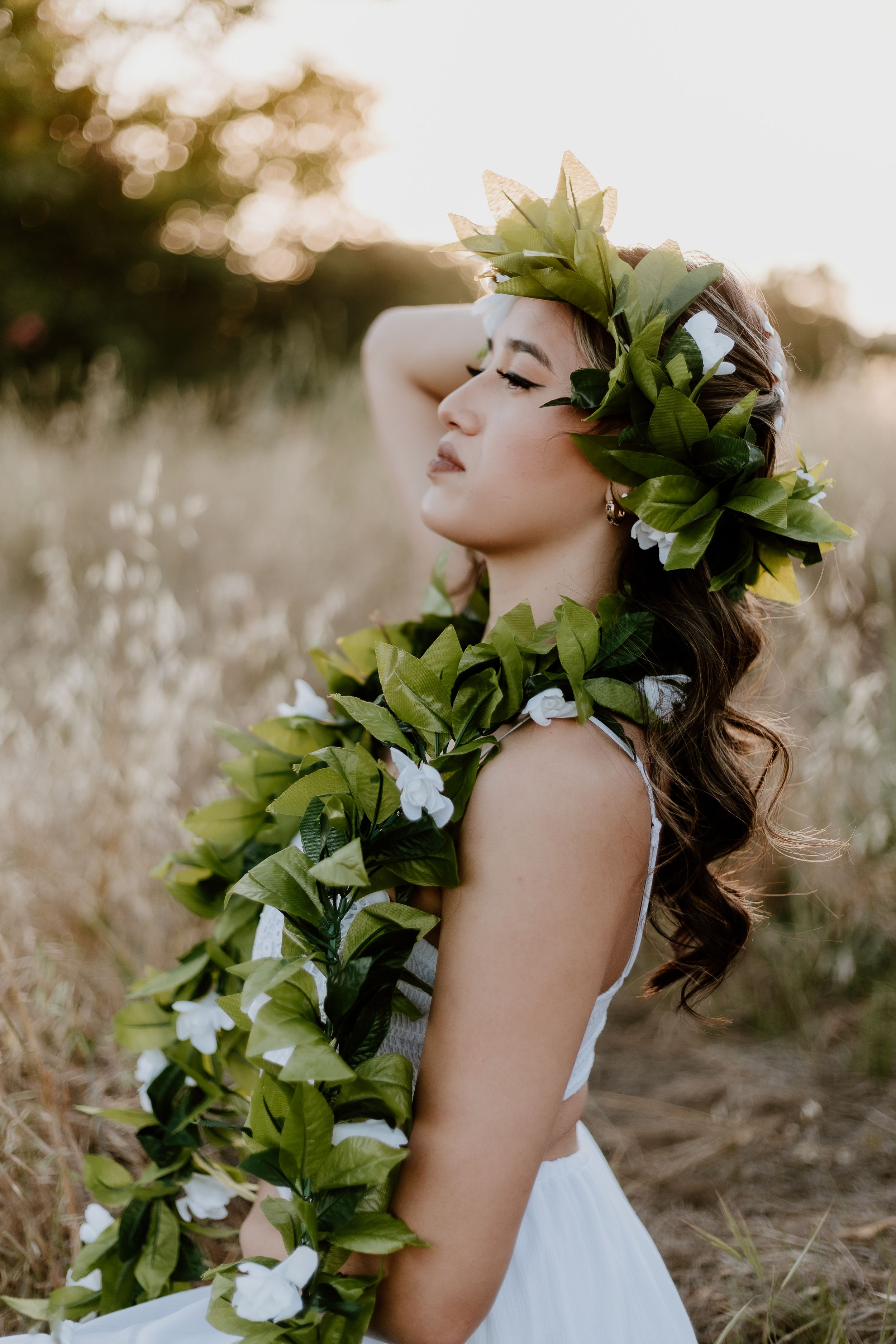 A beautiful woman adorned with a lush green leaf crown and floral lei poses in a golden field.