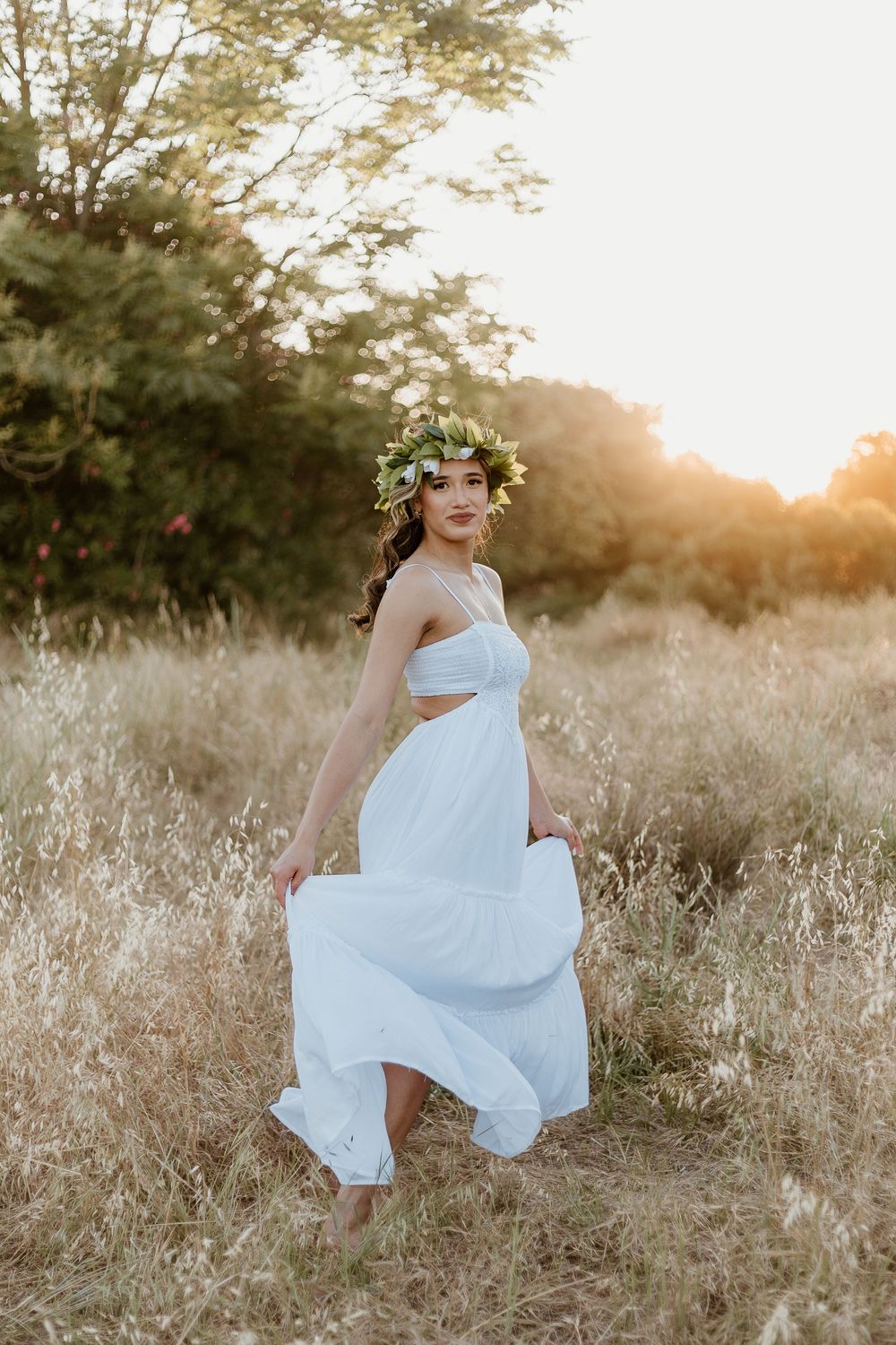 Woman in white dress and floral crown stands in golden field at sunset.