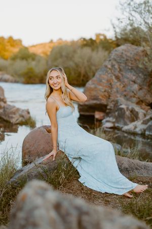 Blonde woman in a light blue maxi dress sitting on rocks by a river at golden hour sunset.