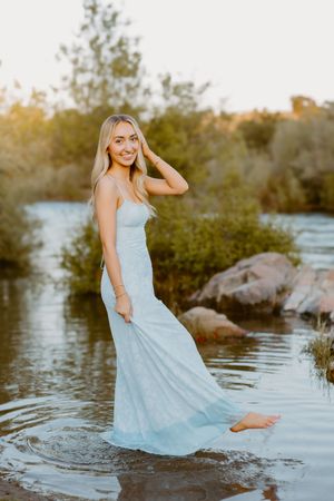 Young blonde woman in light blue dress standing barefoot in a shallow river at golden hour sunset.