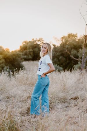 Smiling blonde woman in white tee and wide-leg jeans standing in golden dry grass field at sunset.